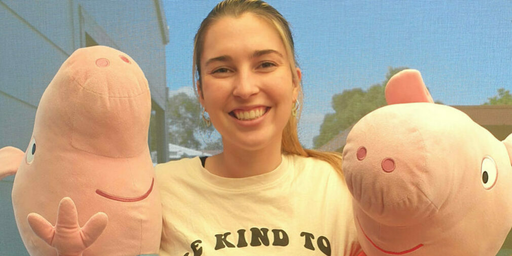 A lady with a big smile, wearing a tshirt that says "be kind to our friends", and hugging large plush Peppa and George Pig toys.