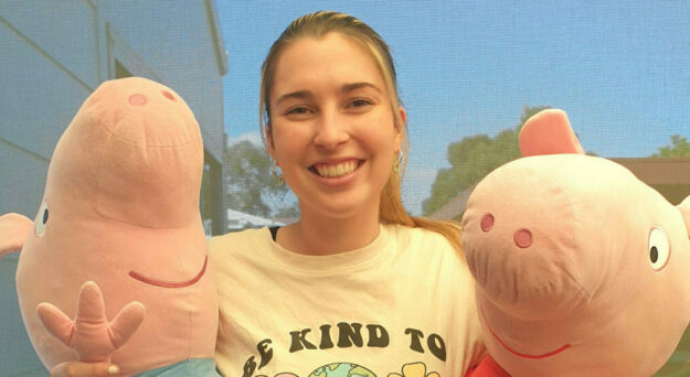 A lady with a big smile, wearing a tshirt that says "be kind to our friends", and hugging large plush Peppa and George Pig toys.